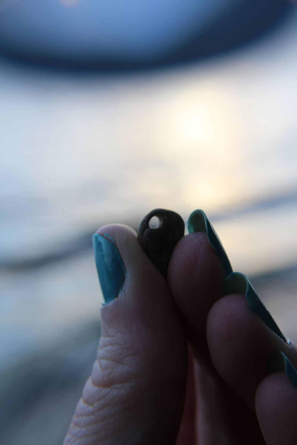 Closeup of Molly’s fingers with blue nail polish holding up a small shell with a very blurred lake in the background 